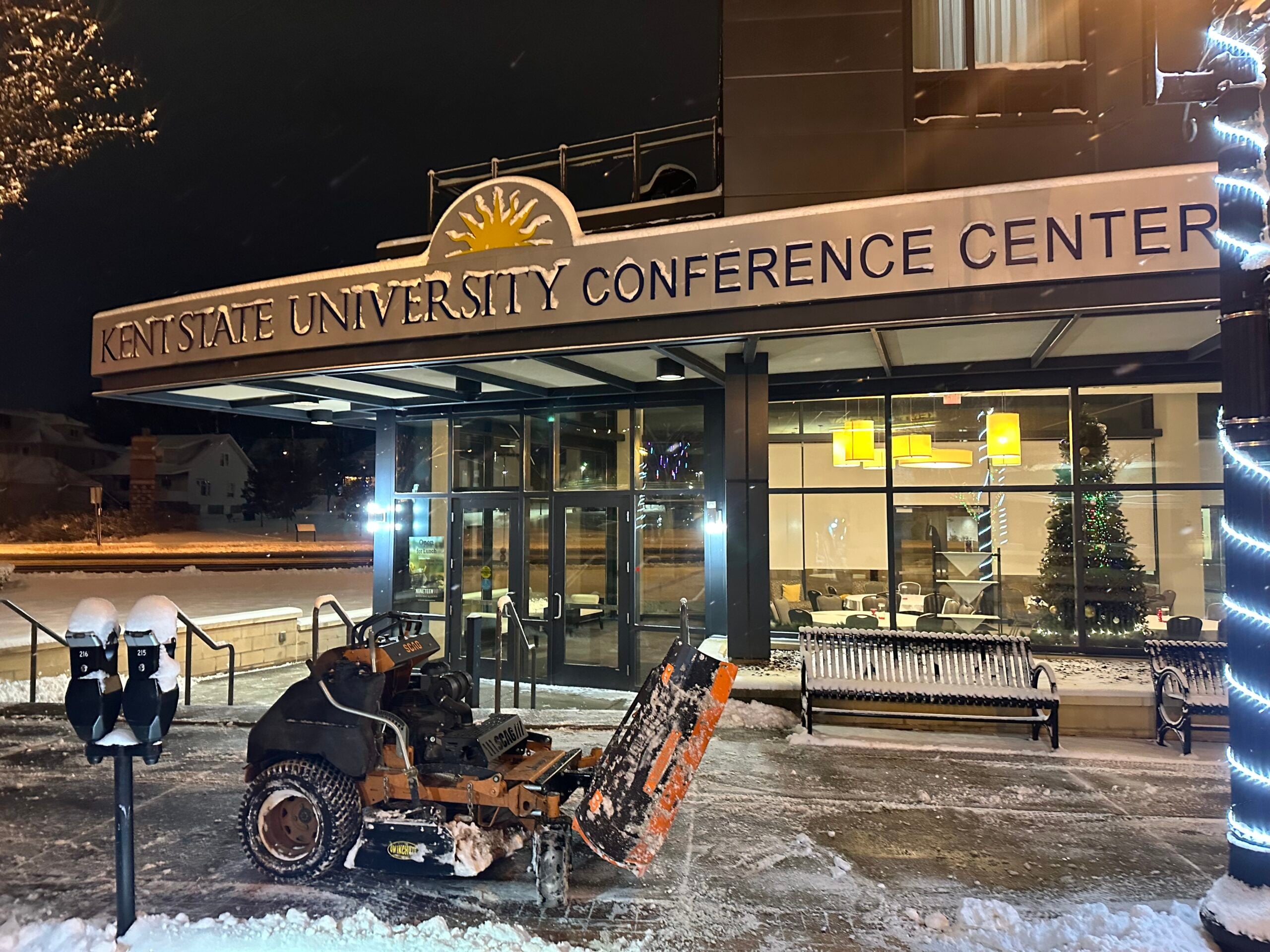 The outside of Kent State University Conference Center has been freshly plowed of snow. On the sidewalk, a small black and orange plow can be seen.