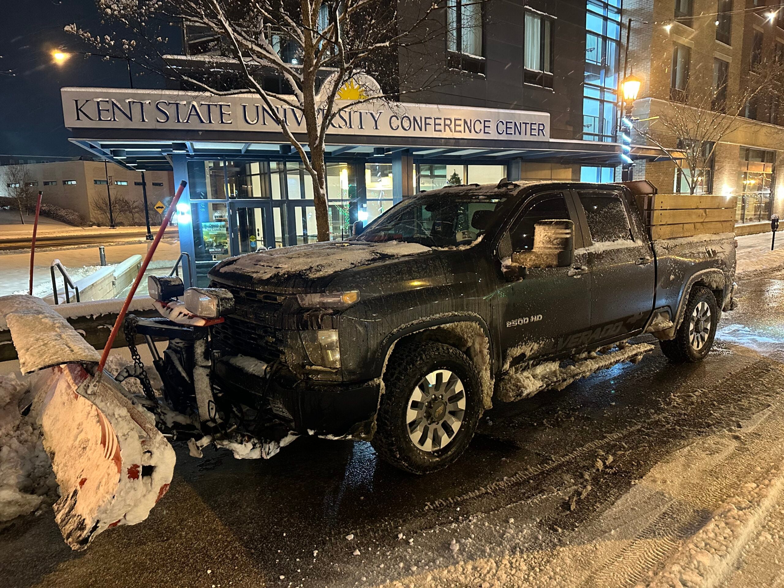 In front of the Conference Center at Kent State, a black truck with a plow on its front can be seen. The streets around it have had snow removal services.