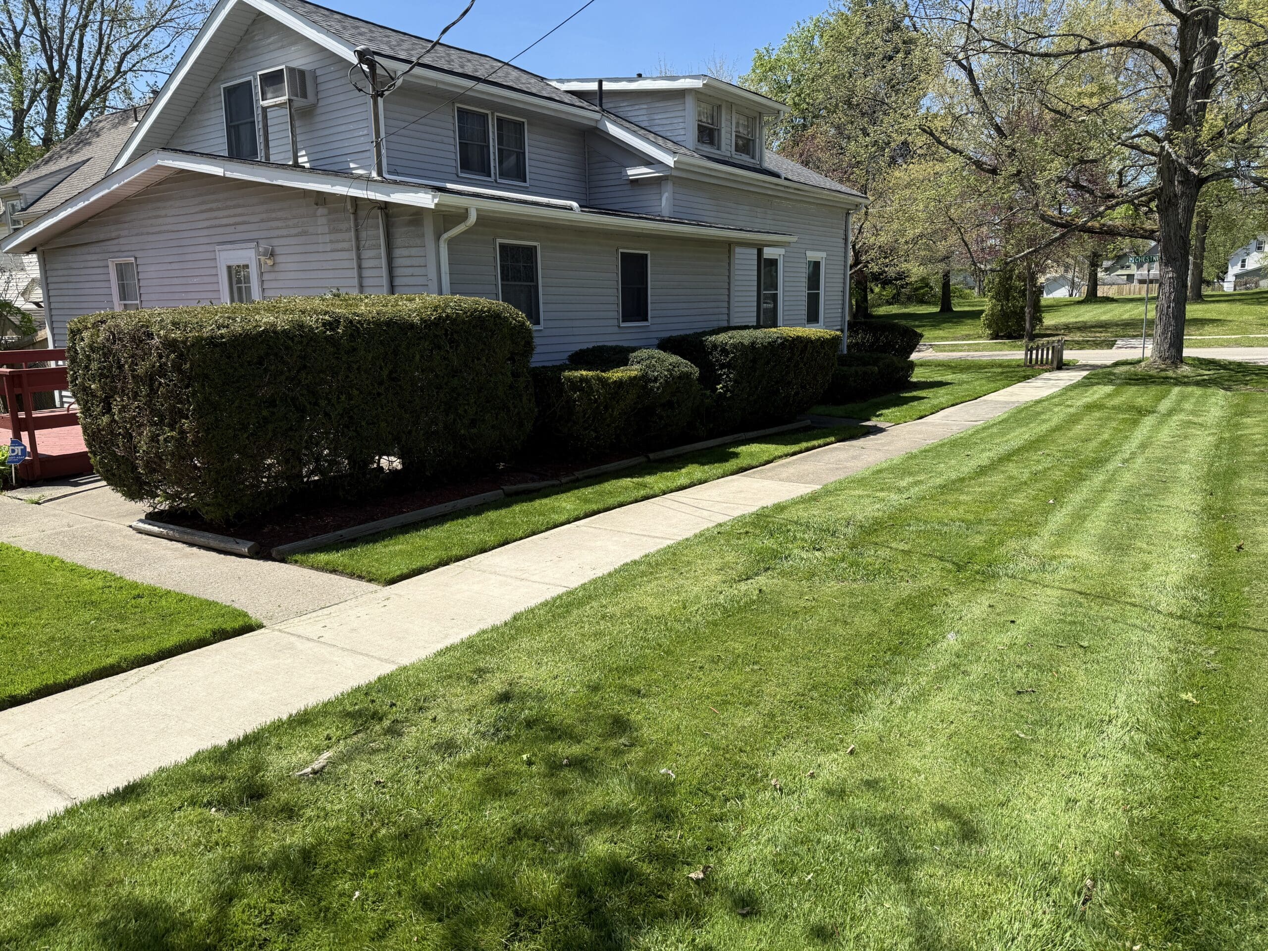 Next to the sidewalk near a grey house, a green lawn shines brilliantly, freshly trimmed.
