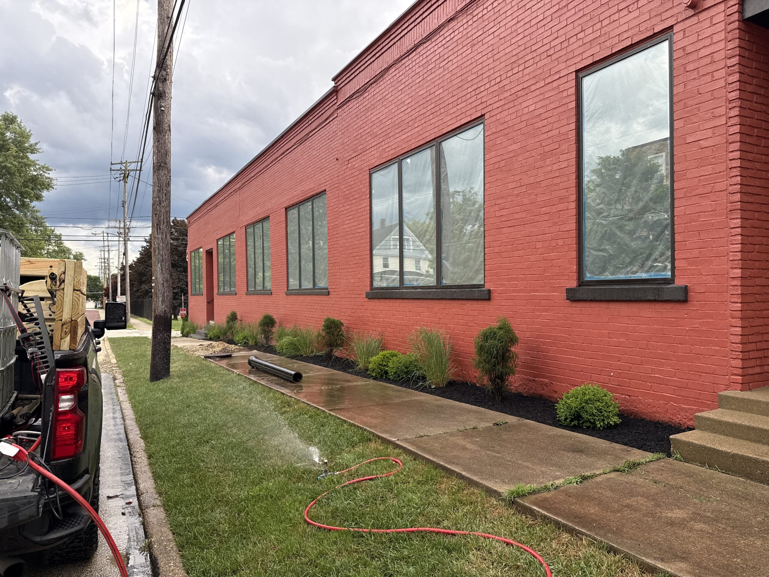 Beside a red building in a town, grass was recently cut near a sidewalk. A sprinkler system, placed by Pro Finish, can be seen watering the grass.